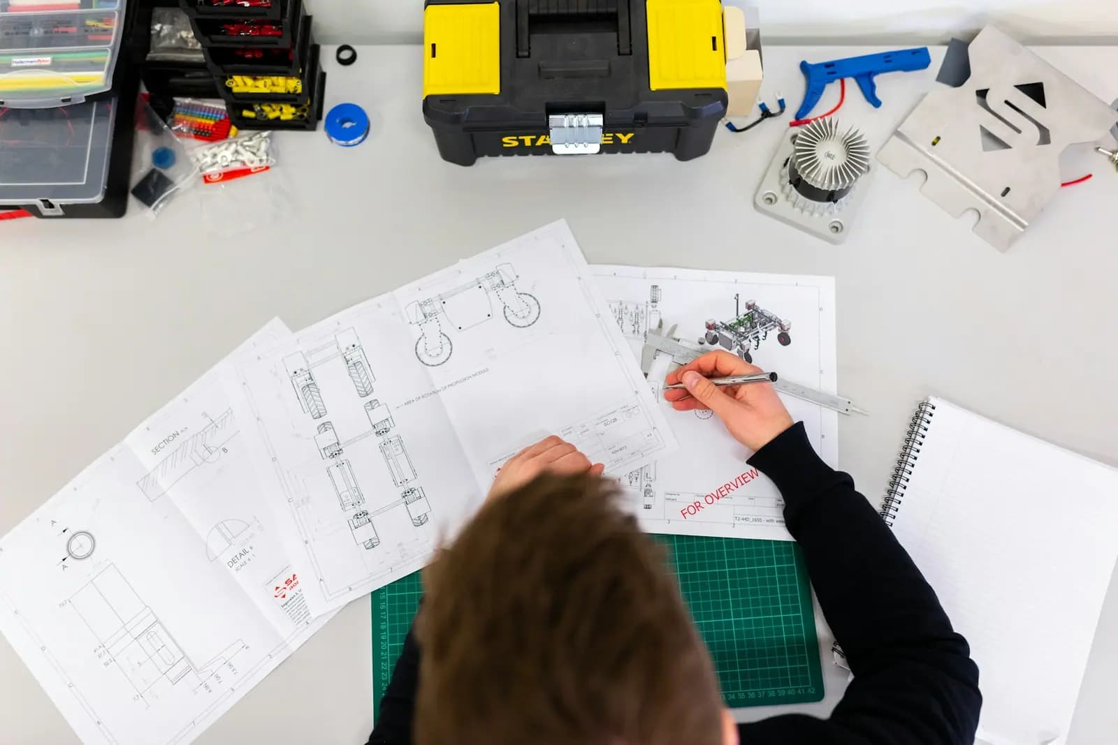 Draufsicht auf ausgebreitete technische Zeichnungen, eine Schieblehre und Bauteile auf einem Arbeitstisch.